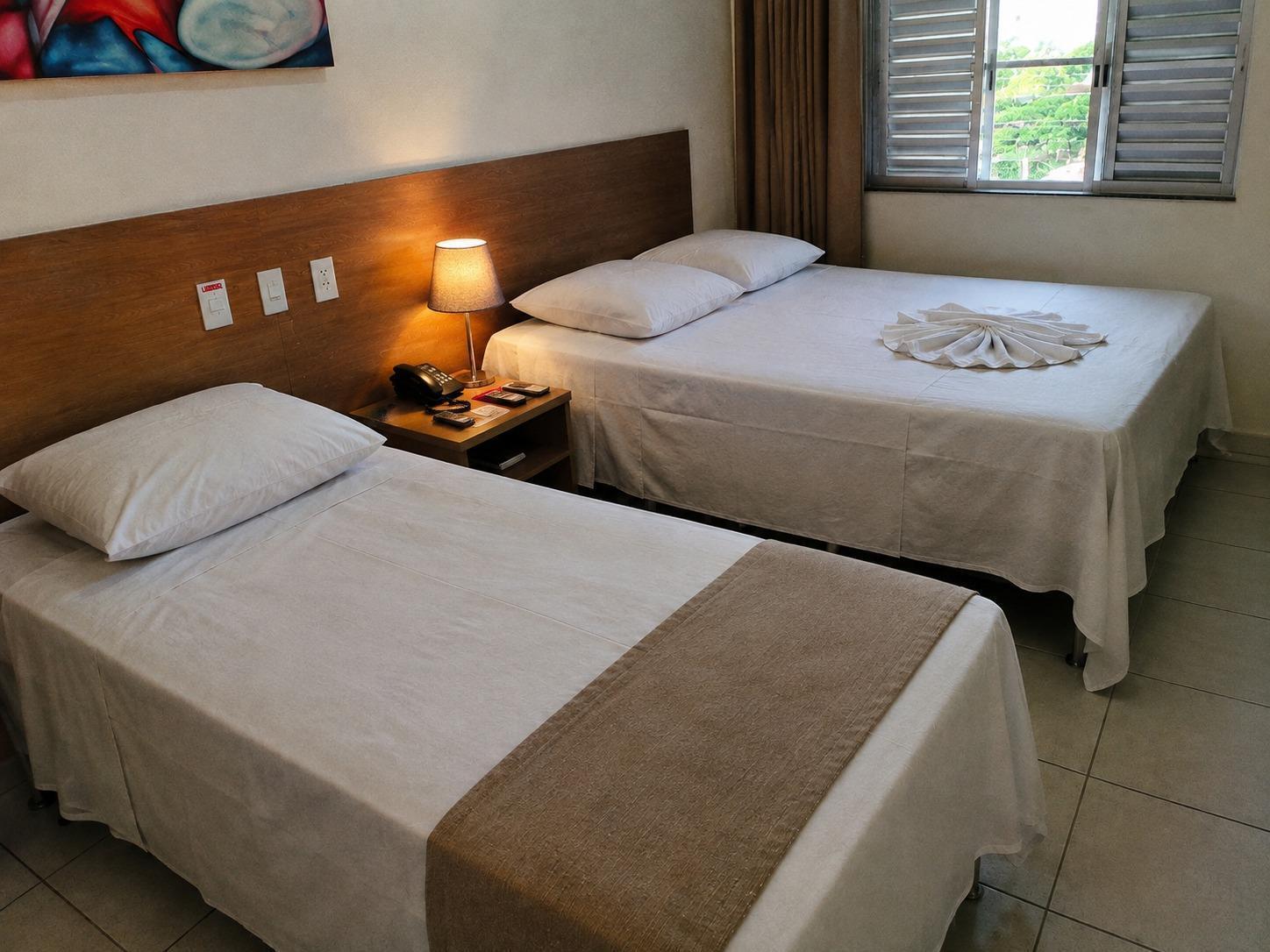Quarto com uma cama de casal e uma cama de solteiro, criado-mudo com abajur e telefone entre elas e janela com cortina deixando entrar luz natural