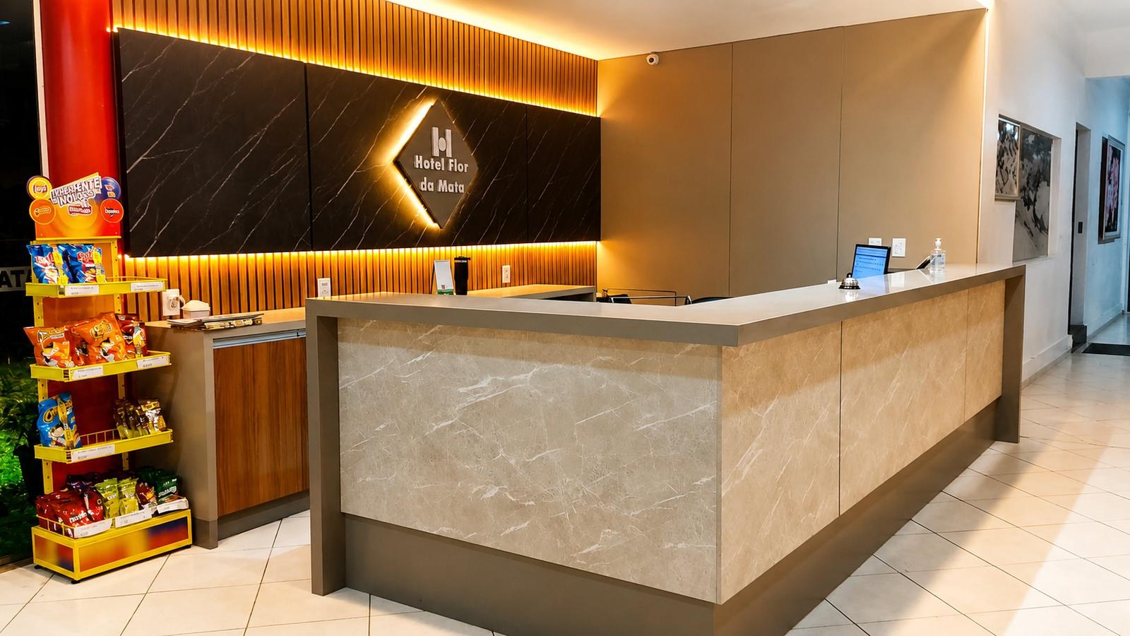 Vista ampla da recepção do Hotel Flor da Mata, com balcão em mármore claro, painel de madeira ripada iluminado, logotipo do hotel e corredor de acesso aos quartos